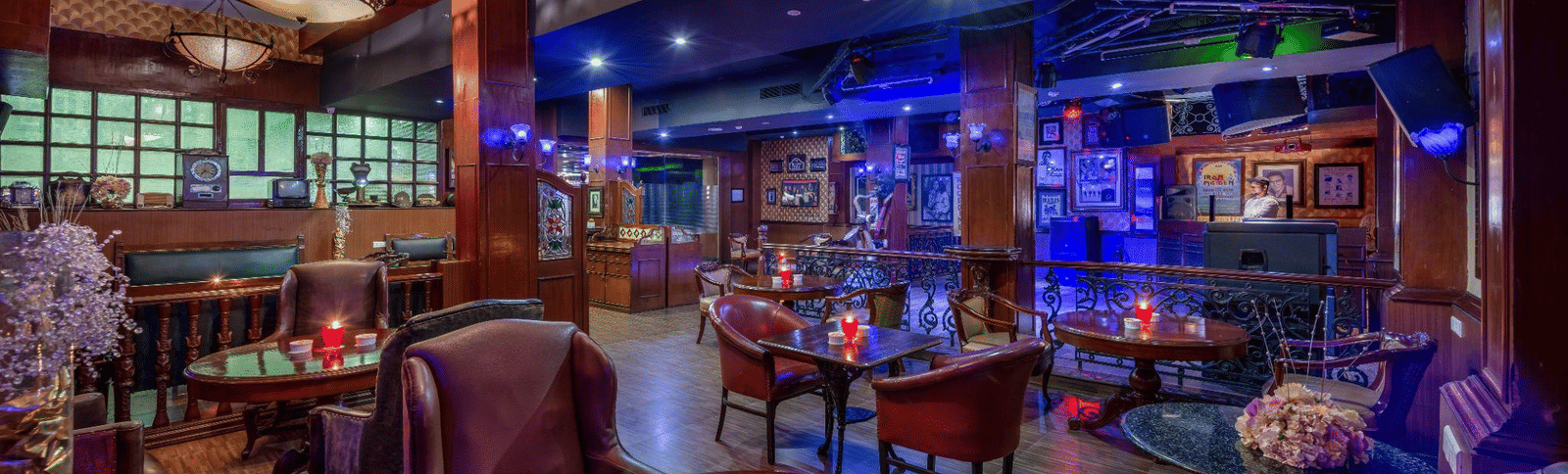 Dark, atmospheric bar or nightclub with red and purple mood lighting, featuring small tables, plush chairs, and a lively vibe