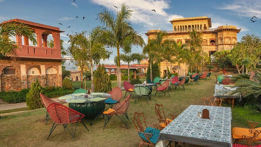 Outdoor seating with colorful chairs and tables is arranged on the lawn of Neemrana's Tijara Fort Palace under a partly cloudy sky.