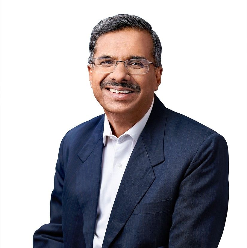 Rajesh Ranjan in a dark blue suit and white shirt, setting on a wooden stool with arms on this knees, smiling looking at camera