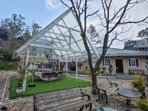 An outdoor seating area with a clear, peaked roof structure, surrounded by gravel and featuring a bare tree, benches, and round tables, with a building in the background.