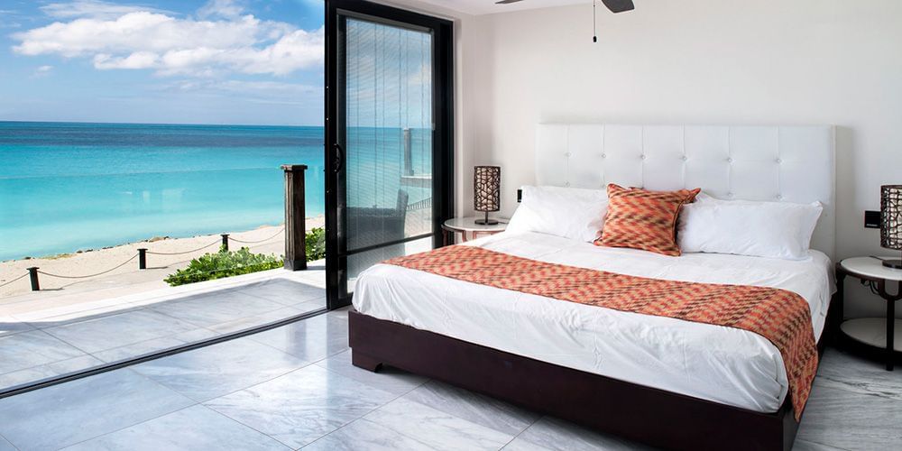 Bright oceanfront bedroom with floor-to-ceiling windows, modern white bed, and direct views of the beach and vibrant blue sea.