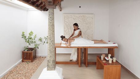 A therapist administering a massage to a person lying on a wooden treatment table in an open-sided room with a stone pillar | Le Pondy