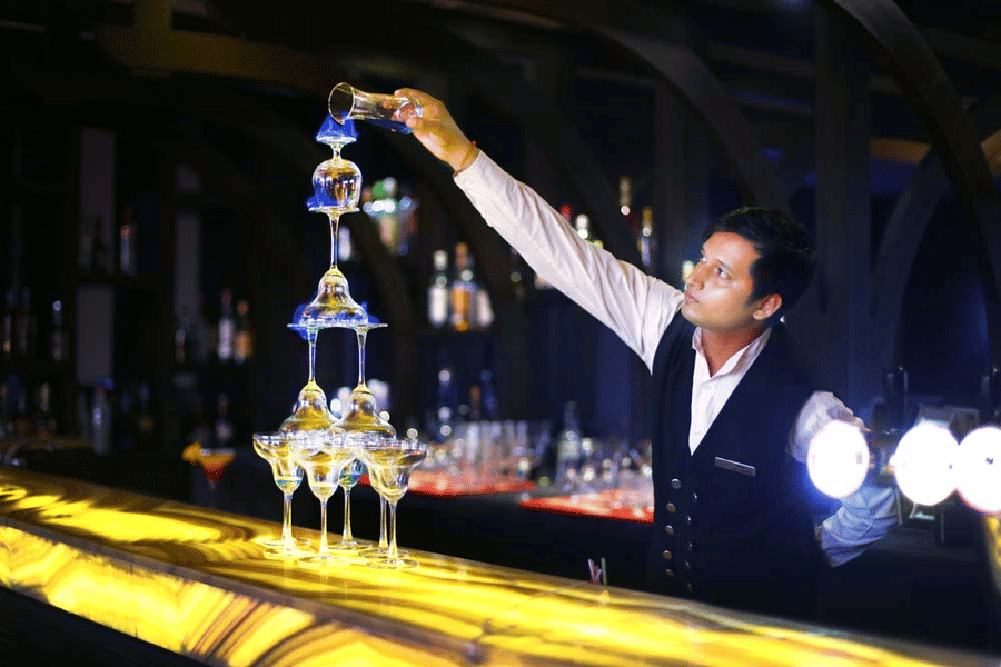 Bartender at The Bristol Hotel, Gurgaon pouring blue liqueur down a tiered champagne glass tower on a backlit bar counter lined with bottles.