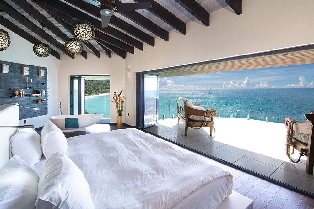 A spacious room with plush white bedding and a large wall to wall window with a view of the ocean.