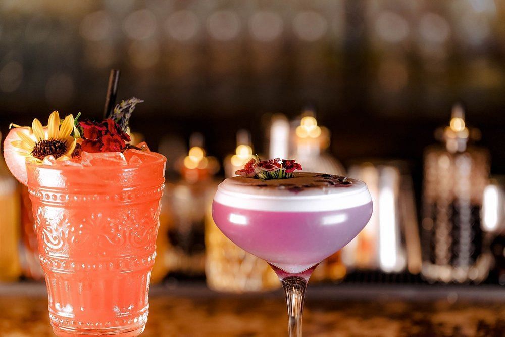 Glass filled with orange beverage topped with a flower accompanied by another glass filled with a lavender purple and pink beverage topped by flowers with a  blurred background.