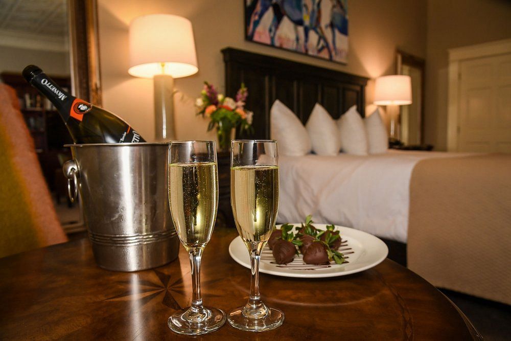 Front view of two champagne glasses filled with champagne , a steel ice bucket and a plate of food on a table in dimly lit room with a neatly made bed and lamps on either side of the bed