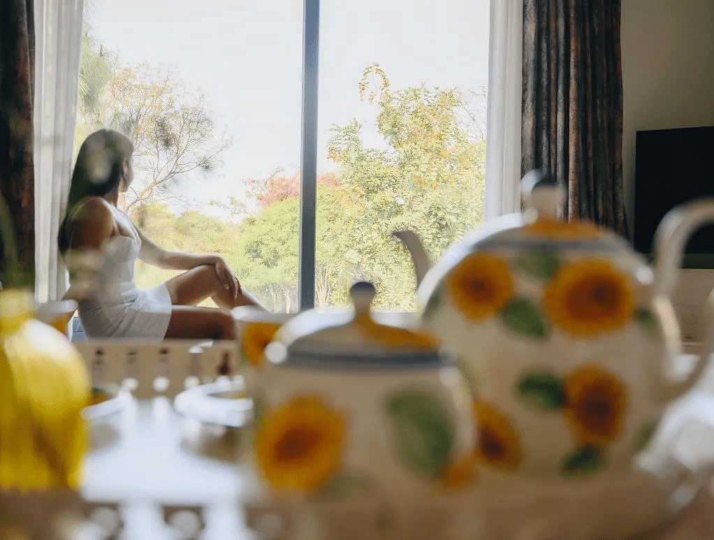 A woman sitting near a sunlit window with a floral teapot and teacups on the table in the foreground at Karma Chalets.