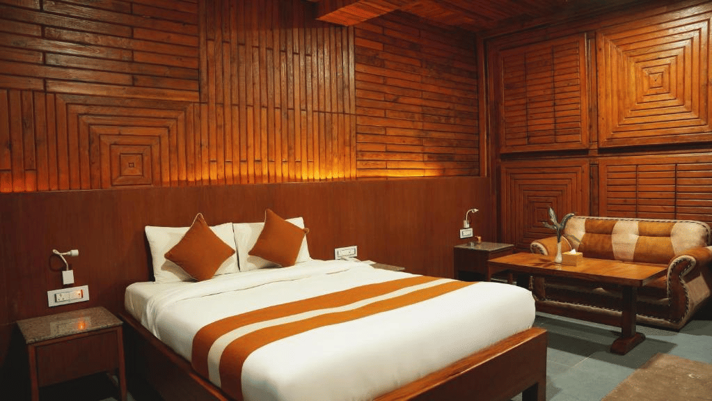 Guest room with wood-panelled walls, a double bed with an orange runner, reading lamps, a sofa and a wooden side table at Ila Comfort, Harkapur.
