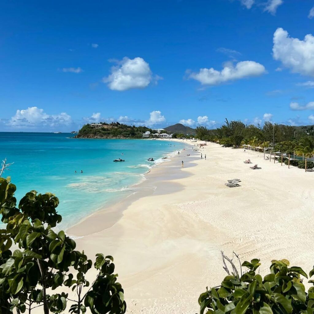 A panoramic view of Ffryes Beach with turquoise waters, white sand, distant hills, and people enjoying the sunny day.