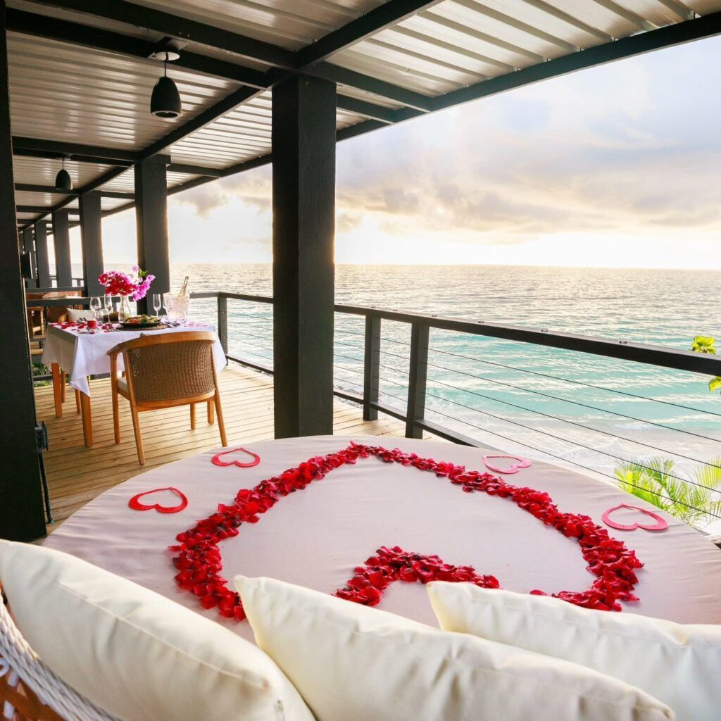 A romantic outdoor dining setup on a balcony overlooking the ocean at sunset, with a heart-shaped rose petal arrangement on the bed.