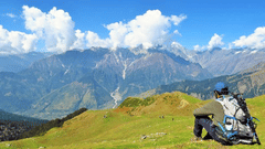 A picture of a man sitting on a hilly terrain with Trekking equipment overlooking the majestic green mountains - Ramgarh Bungalows, Nainital,