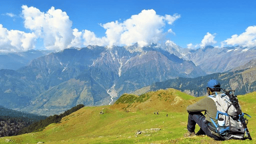 A picture of a man sitting on a hilly terrain with Trekking equipment overlooking the majestic green mountains - Ramgarh Bungalows, Nainital,
