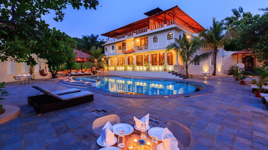 The brightly lit facade of the resort building during the evening with the outdoor swimming pool in the foreground, with sun loungers and a small dining table decorated with candles - Golden Tulip Goa Mandrem