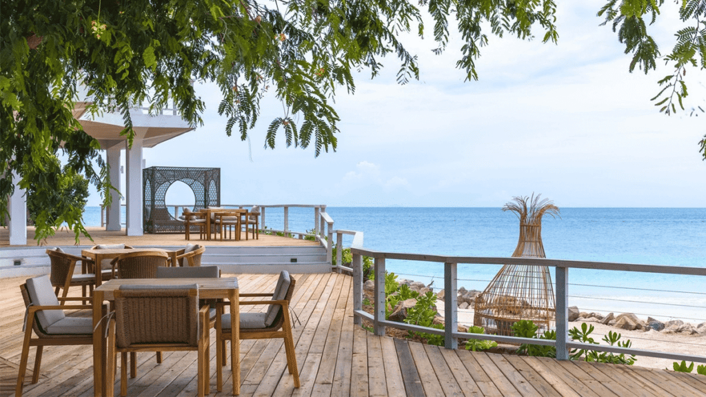 An outdoor dining area on a wooden deck offers panoramic views of the ocean, framed by lush trees.
