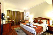 Standard Room at The Bristol Hotel, Gurgaon with a king-size bed, rust-coloured runner, a towel swan decoration, a wooden desk, an armchair, and curtains.