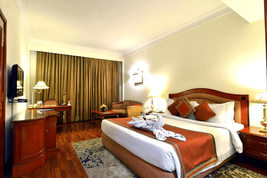 Standard Room at The Bristol Hotel, Gurgaon with a king-size bed, rust-coloured runner, a towel swan decoration, a wooden desk, an armchair, and curtains.