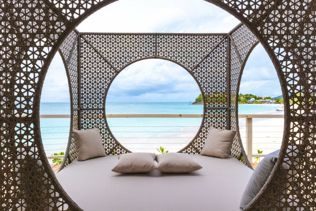 A circular woven cabana with cushions looking out onto a white sand beach and a clear turquoise ocean under a cloudy sky.