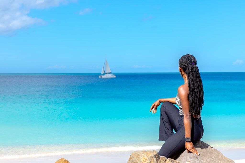 A woman sitting on a rock in the beach side, facing the blue sea.
