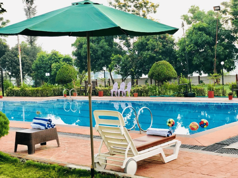 An outdoor swimming pool is bordered by a paved deck, with white sun loungers and a large green umbrella at VITS Kamats Resort, Silvassa