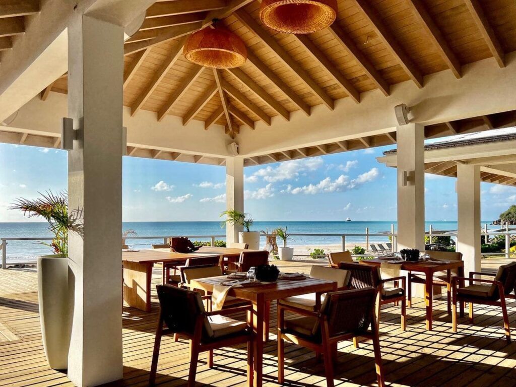 Open-air seaside restaurant with wooden furniture and a high thatched roof, overlooking a sunny ocean view under a clear blue sky.