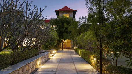A tiled pathway with exterior lights illuminating it, surrounded by well-maintained gardens during the evening hours at Anandam Jungle Retreat Resort, Udaipur.