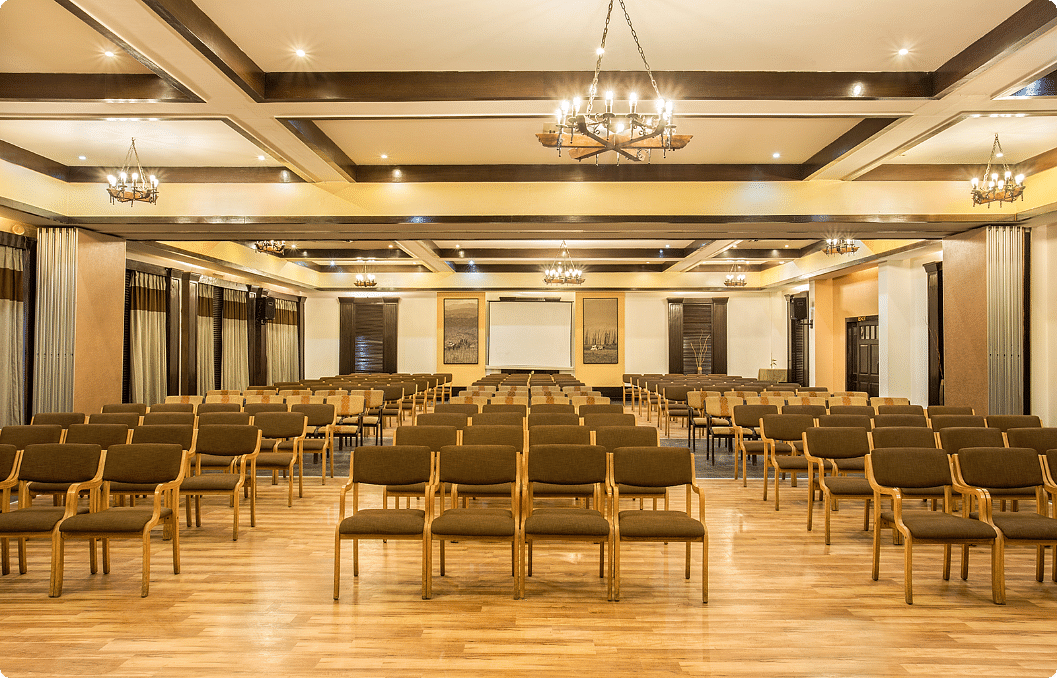An image of a conference hall with a set of table and chairs in each corner and a projector screen at the front of the room - The Riverview Retreat, Corbett