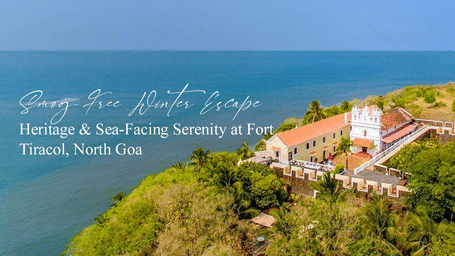 A scenic aerial view of a large heritage building with white walls and red roof situated on a lush green hilltop, overlooking the sea in North Goa. Text on the image reads: Smog-Free Winter Escape: Heritage & Sea-Facing Serenity at Fort Tiracol, North Goa.