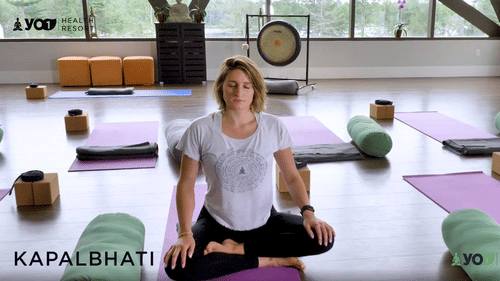 A woman sits cross-legged on a yoga mat in a studio with a gong in the background, facing forward. The text KAPALBHATI is visible at YO1 Longevity & Health Resorts