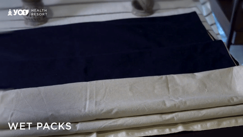A flat, dark blue cloth lies on top of a folded stack of light-colored, possibly wet, sheets on a surface, with WET PACKS text visible at YO1 Longevity & Health Resorts