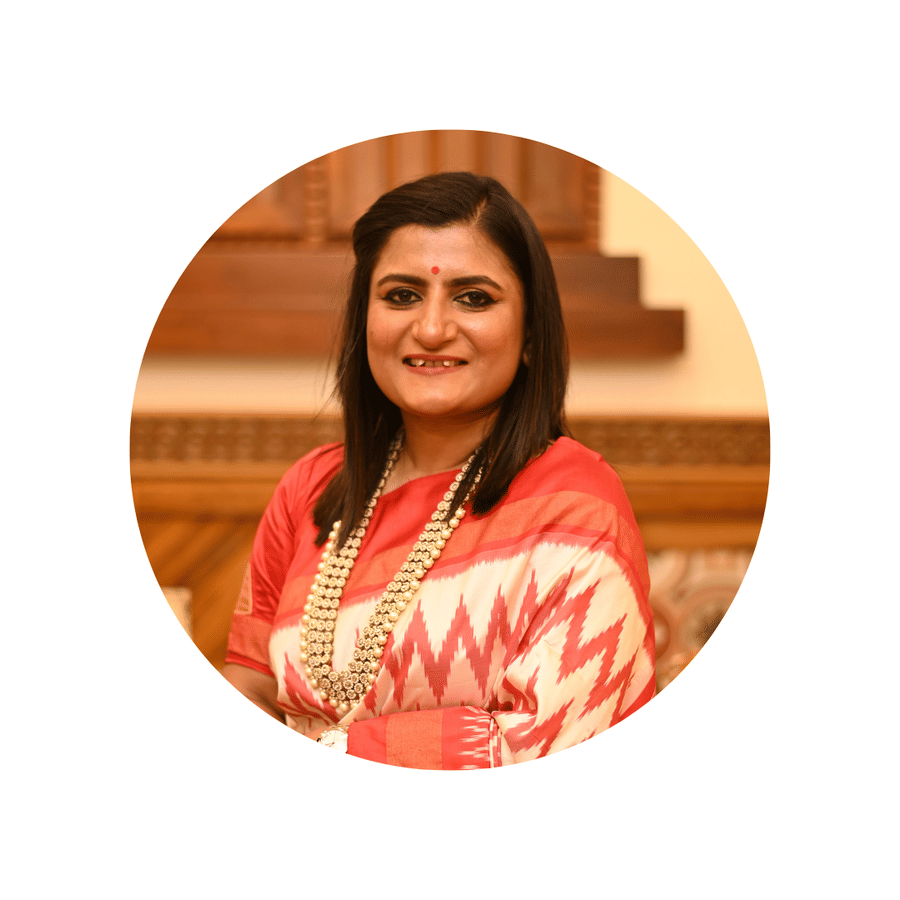 Portrait of  Vidhi Vikram Kamat, Director of VITS Kamat Group, smiling in a red and white patterned traditional outfit, standing indoors with warm wooden decor.