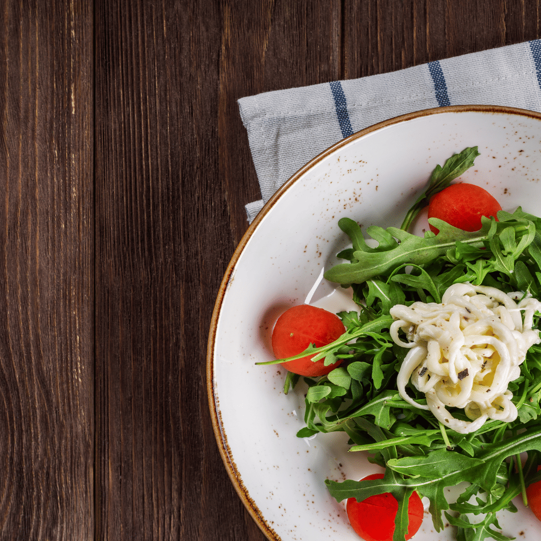 Fresh arugula salad with cherry tomatoes and soft cheese served on a white plate over a rustic wooden table.