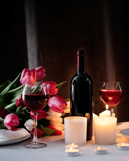 A red wine bottle with 2 filled glasses, tulips and lit candles arranged on a table, creating an intimate candlelight dinner setting.