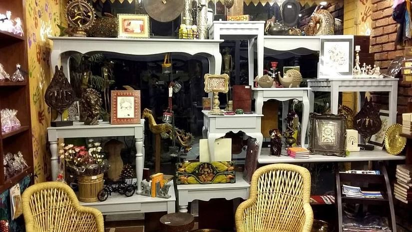  An eclectic room filled with antique furniture, decorative items, and unique artifacts. Two wicker chairs sit in the foreground, facing a collection of mirrored dressers and shelves adorned with lamps, sculptures, and various curios.