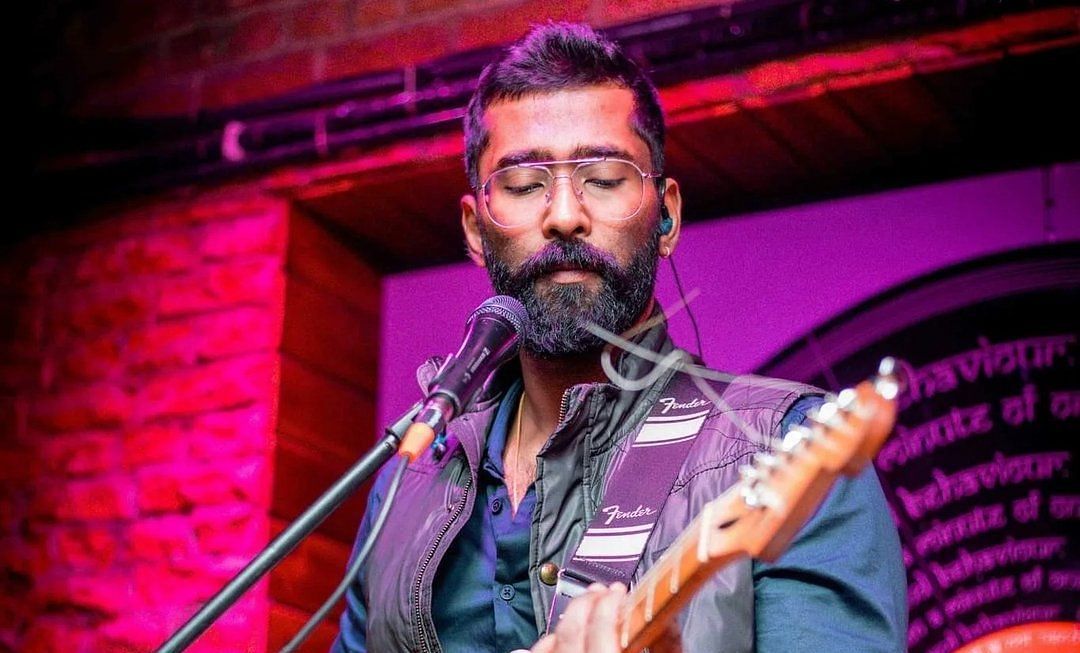 A bearded musician wearing glasses and a stylish vest passionately plays a white electric guitar on stage. 
