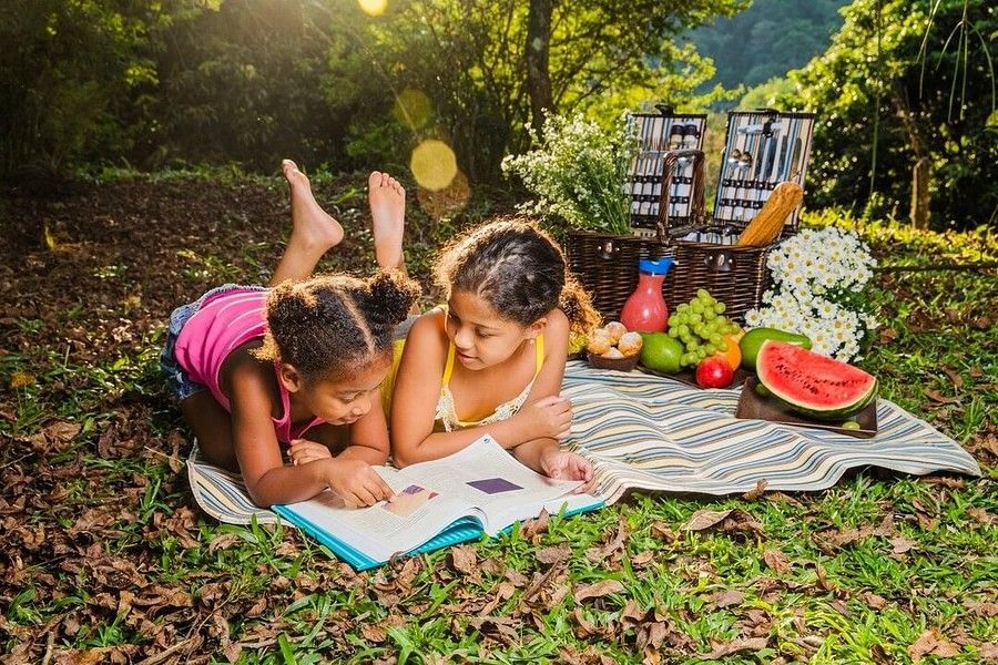 Two girls lying under the sun on a mat with picnic spread on the side and smiling while reading a book