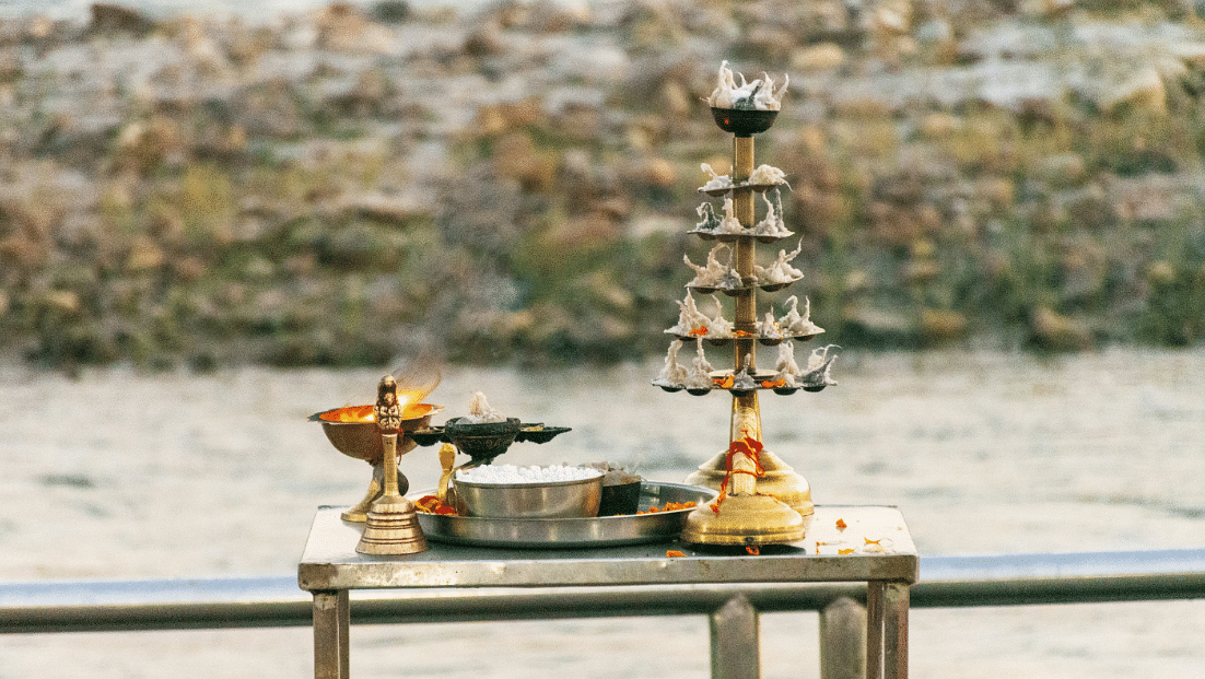 Traditional Indian puja setup on a riverside platform, featuring brass lamps, a kalash, and ritual items.