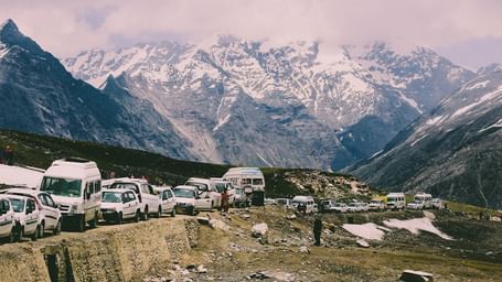 Snow-dusted mountain pass with traffic and parked vehicles, set against a dramatic backdrop of rugged Himalayan.