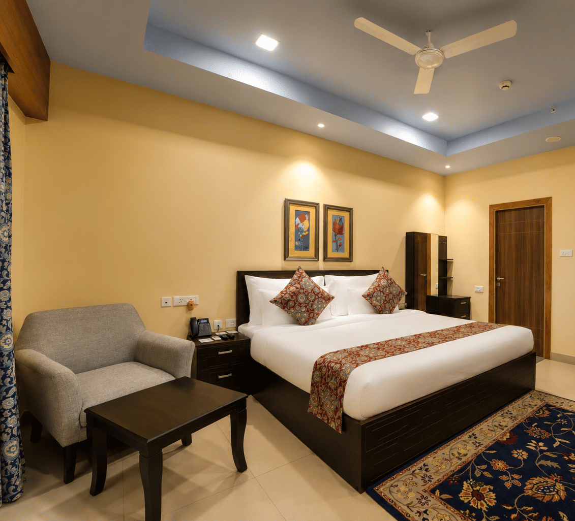 Deluxe suite bedroom with a king bed, grey armchair, coffee table, dark wooden wardrobes and a patterned rug at MAYFAIR World Cup Village, Rourkela.
