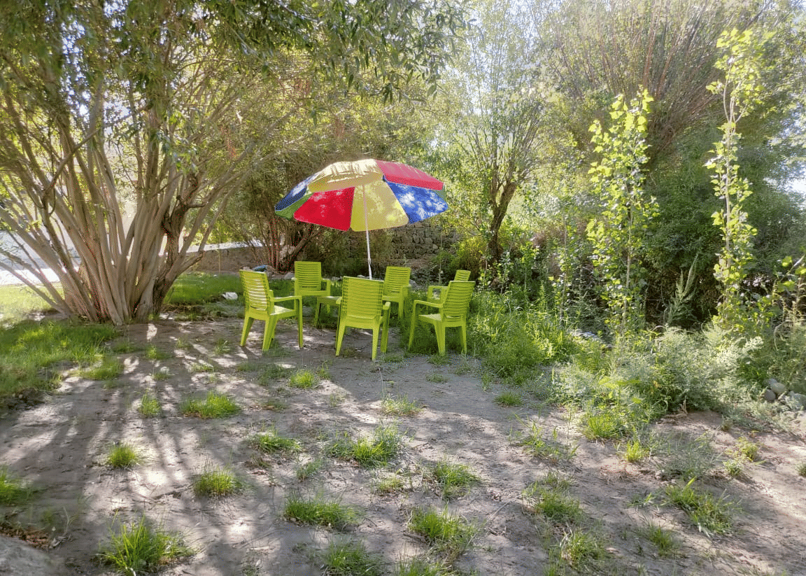 A shaded outdoor seating area with green chairs and a multi-coloured parasol set amongst trees at Indus Serenity Retreat.