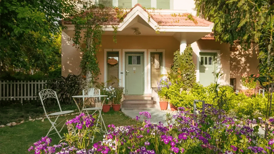 Entrance to Karma Chalets showing a building with red-tiled roof, green door, garden chairs, flowering plants, and trees.