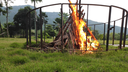 Large bonfire burning in the middle of a grassy field with trees and mountains in the background, set for an outdoor event - Black Thunder, Coimbatore