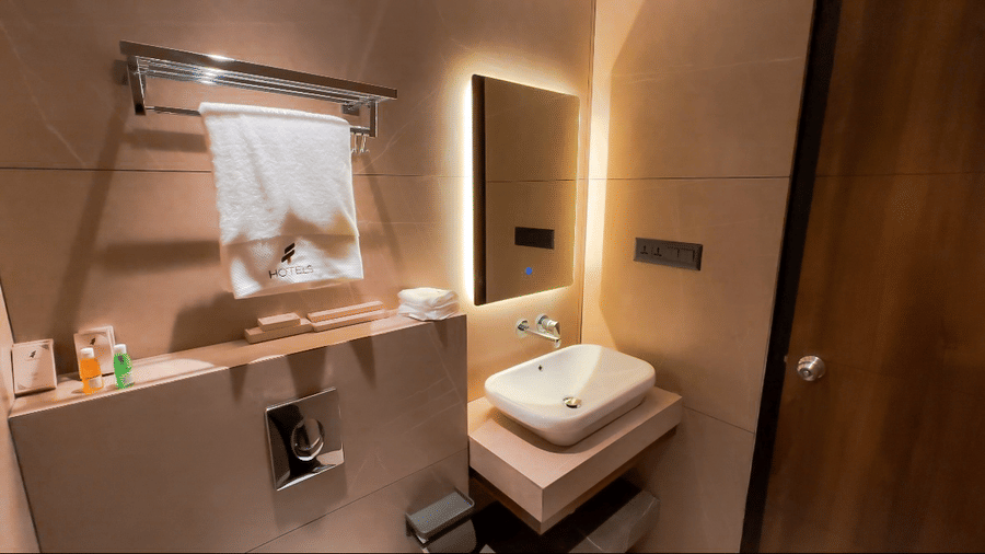 A compact bathroom area with a wash basin and a small square backlit mirror above it at F Hotels, Bengaluru.