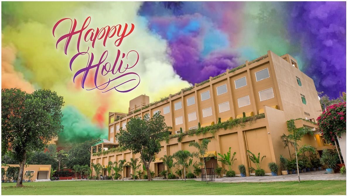 Low angle view of The Lalita Grand Mathura - Vrindavan building surrounded by trees, with a colorful sky in the background and Happy holi written on the top.