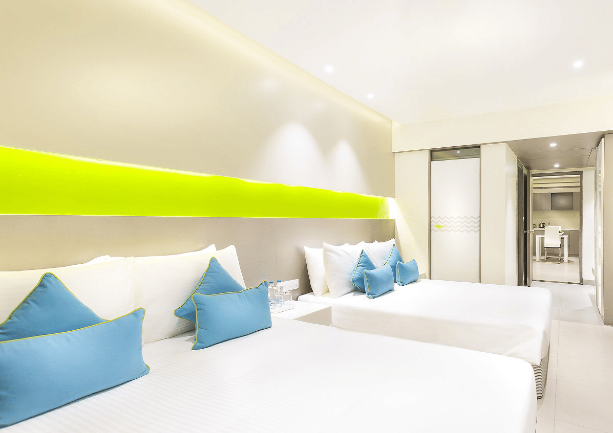 A spacious suite at Zibe Coimbatore by GRT Hotels featuring two large beds with blue cushions, a wall-mounted TV positioned opposite the beds and a headboard stretching across the wall.