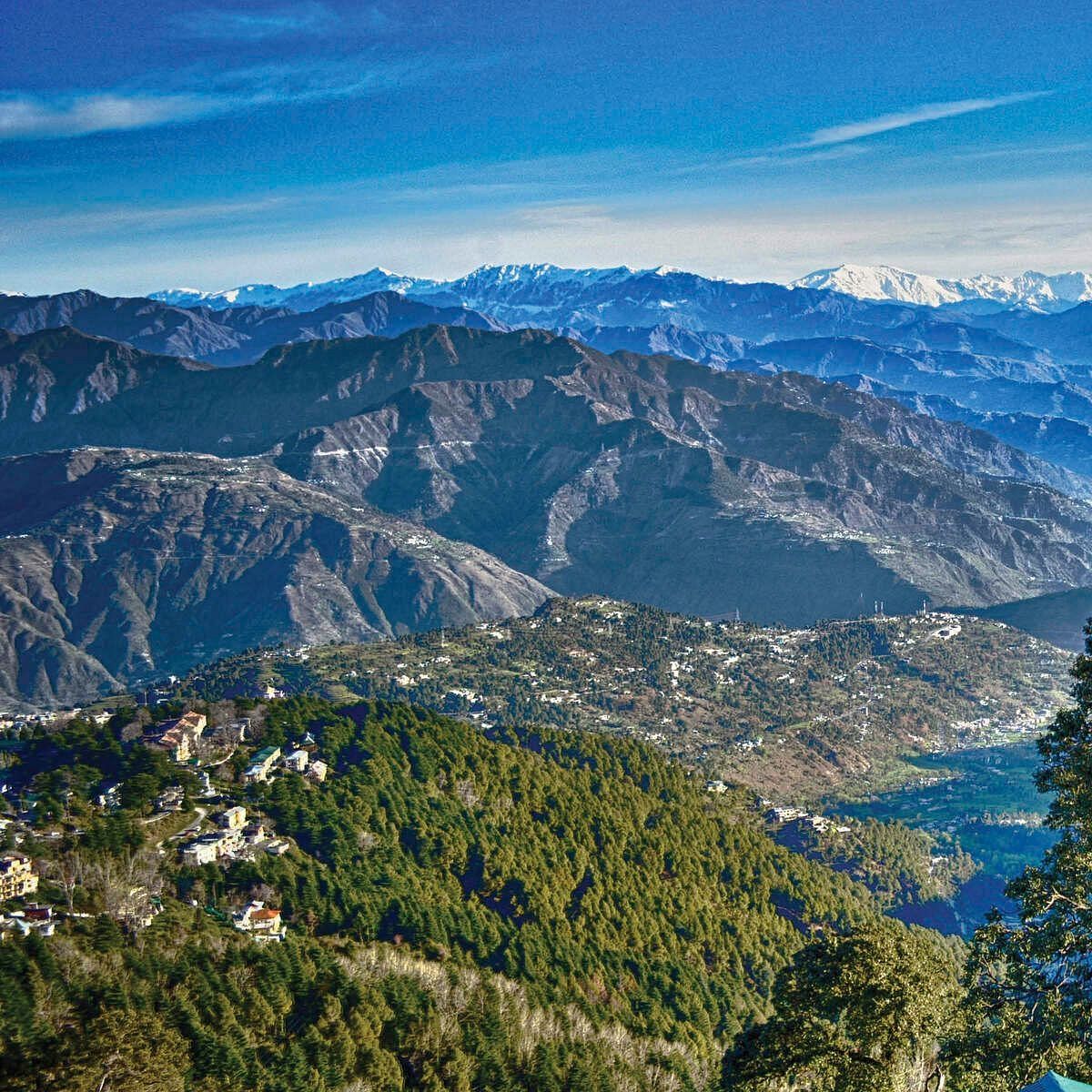 A scenic view of Dalhousie with lush green hills, scattered houses, and snow-capped mountains under a clear blue sky.