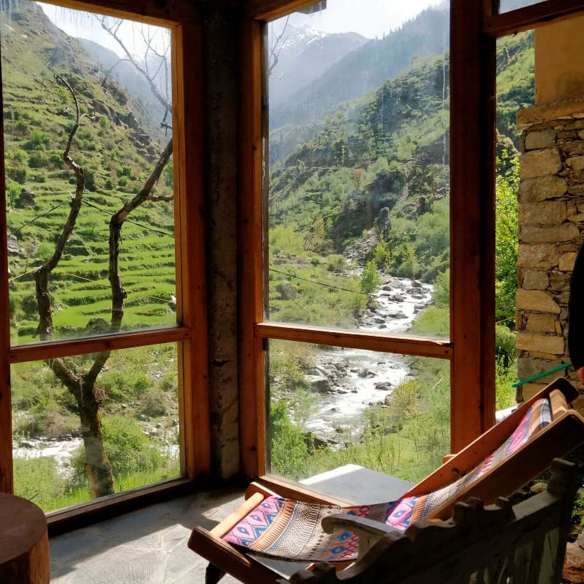 A scenic view of a lush green valley and stream through a wooden window, with a wooden chair.