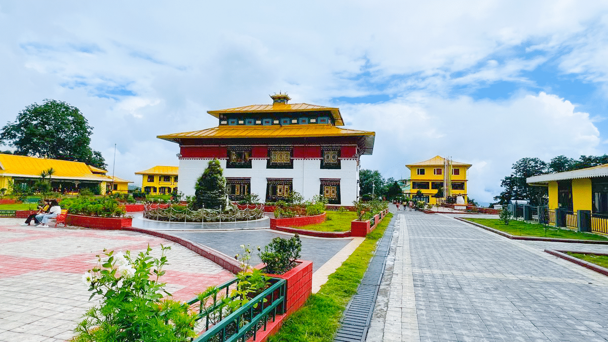 Exterior view of Tsuklakhang Monastery with vibrant yellow and red buildings, landscaped gardens, and a paved walkway under a bright blue sky.