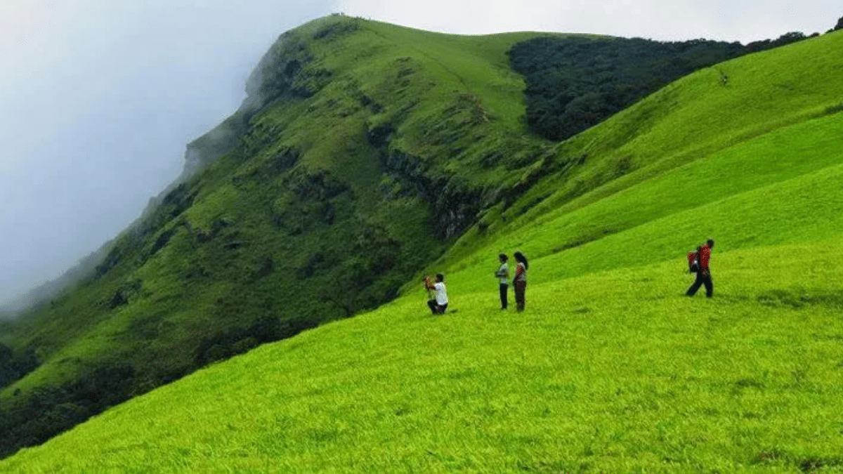 Green hillside trek in Coorg with hikers walking along a rolling ridge trail surrounded by mist and grasslands.