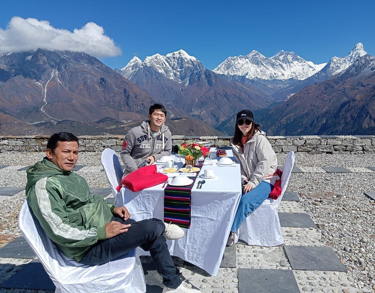 Group of people enjoying a meal at a table set outdoors with a backdrop of snow-capped mountains 