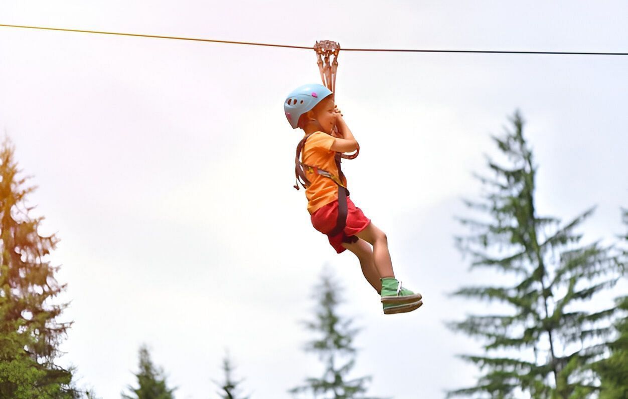 A person ziplining and moving across a backdrop of green trees and a cloudy sky.